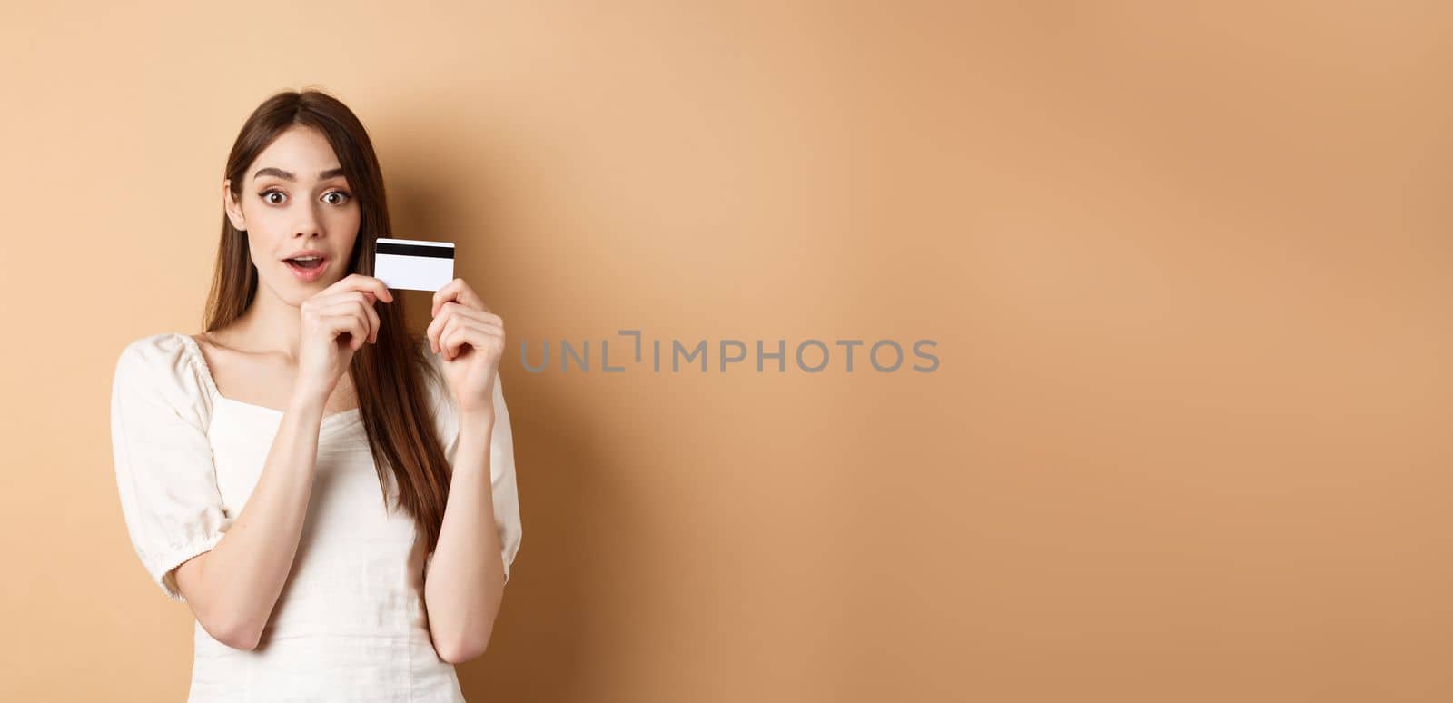 Excited young woman showing plastic credit card and look amazed at camera, standing on beige background.