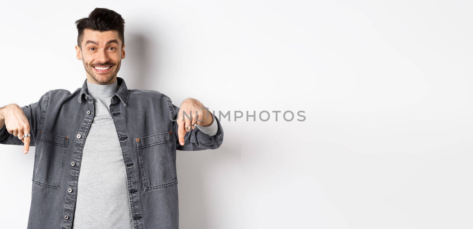 Happy caucasian man with moustache pointing fingers down, smiling and showing special deal, inviting to look here, standing on white background.