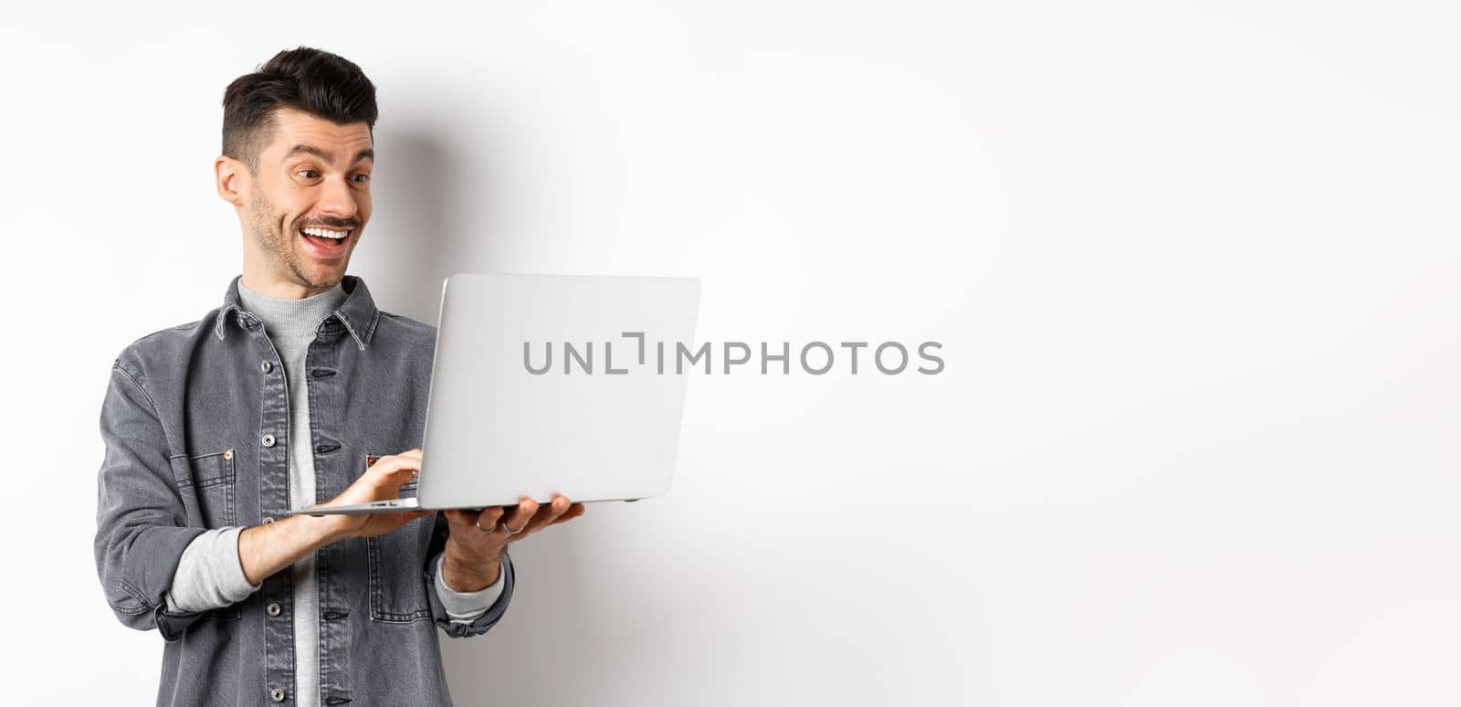 Man watching funny video on laptop, laughing at computer screen and smiling joyful, standing on white background.