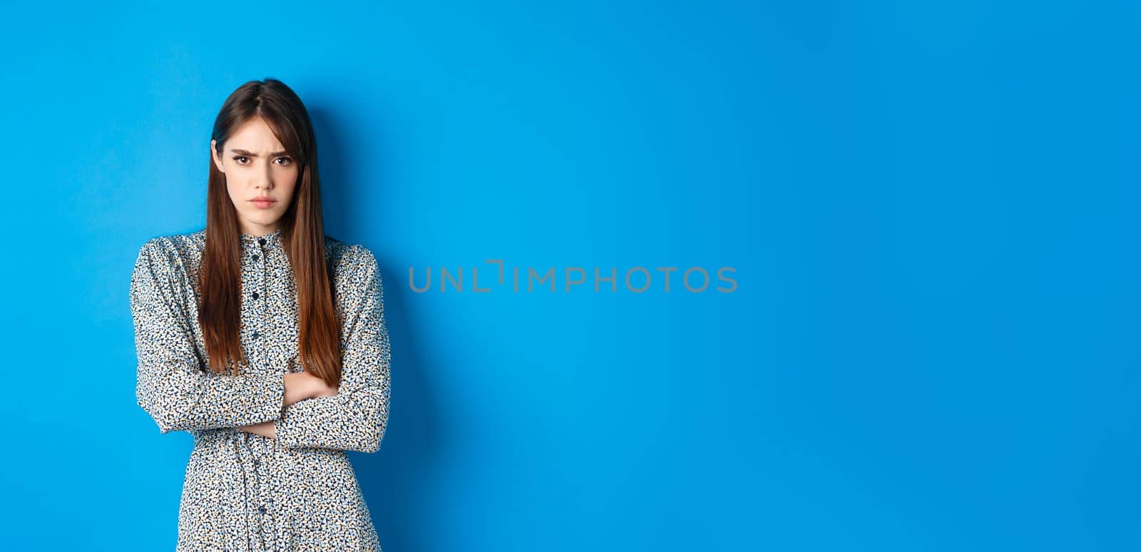 Angry frowning woman in dress, cross arms on chest and feel offended about something unfair, standing upset against blue background.