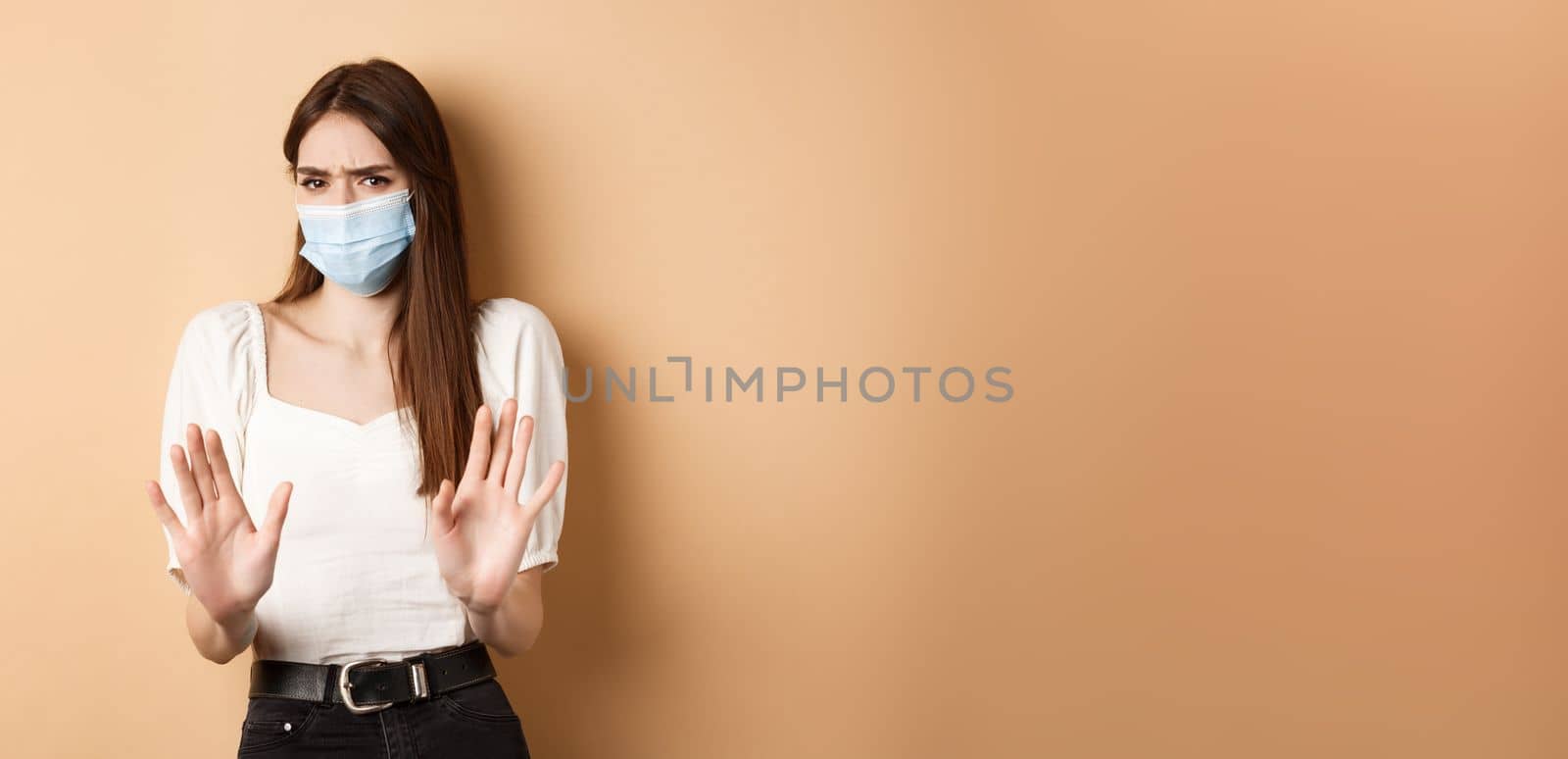 Pandemic and healthcare concept. Keep distance. Young woman in medical mask stretch out hand in stop sign, telling to stay away and frowning disgusted, beige background.