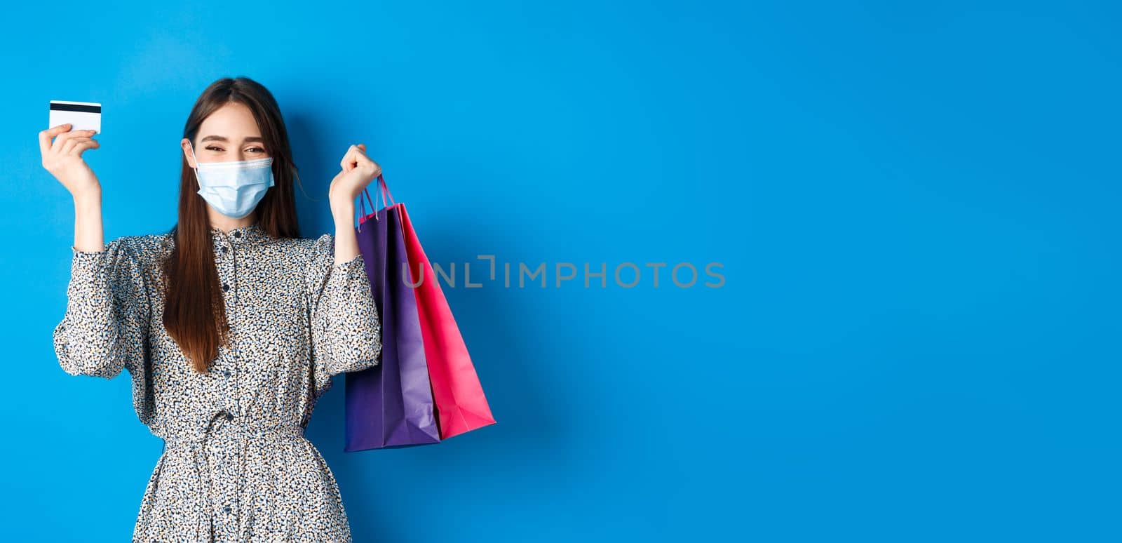 Covid-19, pandemic and lifestyle concept. Pretty female model in medical mask, rejoicing over shopping discounts, holding plastic credit card and bags, standing happy on blue background.