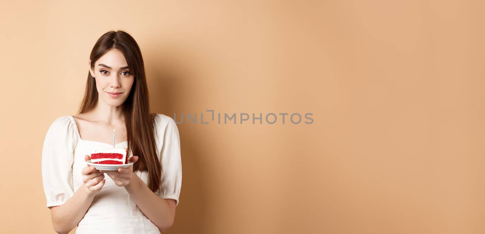 Beautiful birthday girl holding cake with candle, making wish, celebrating b-day on beige background.