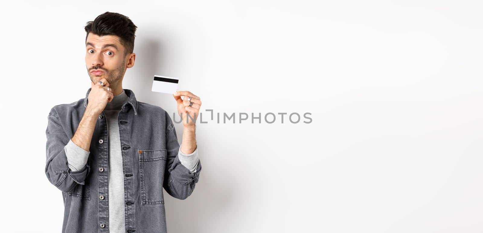 Thoughtful guy holding plastic credit card and thinking, looking intrigued at camera, touching chin while making choice, standing on white background.