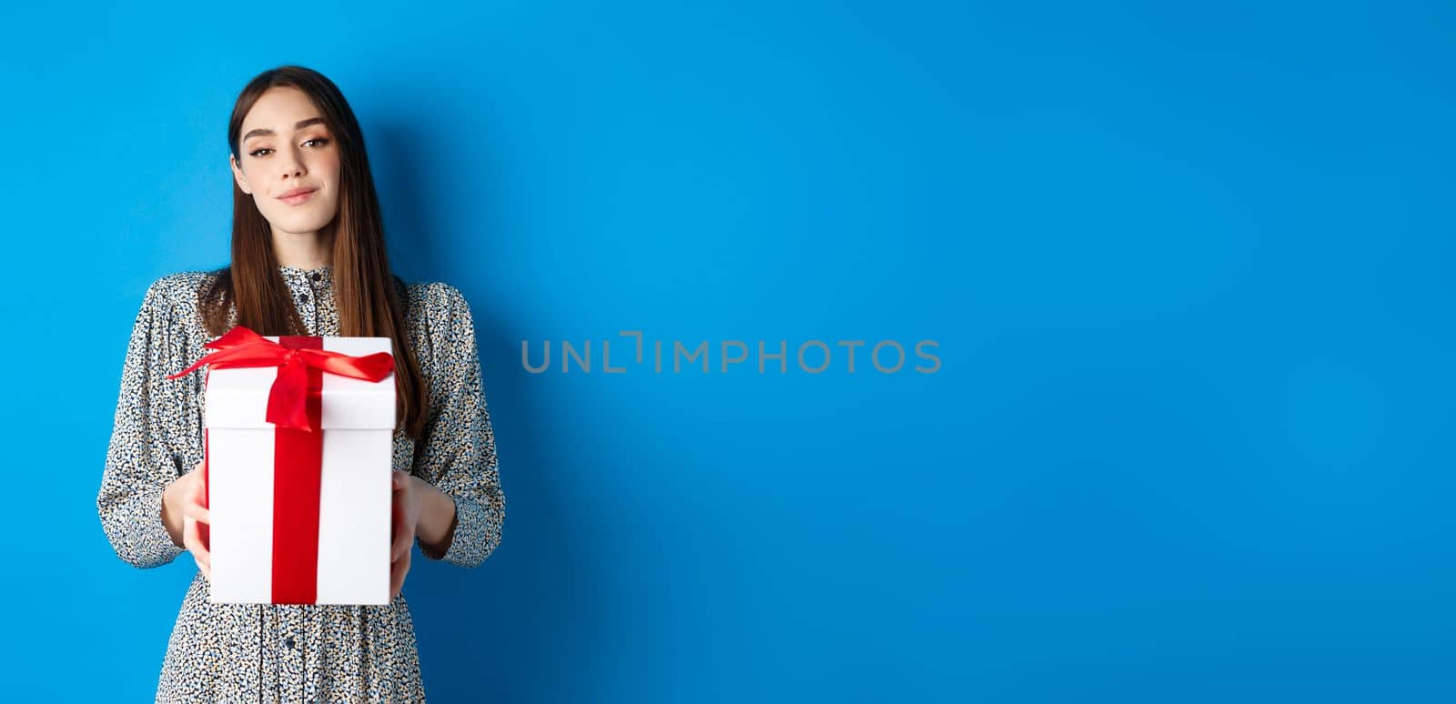 Valentines day. Romantic girlfriend bring gift and smile at camera, standing in trendy dress on blue background by Benzoix