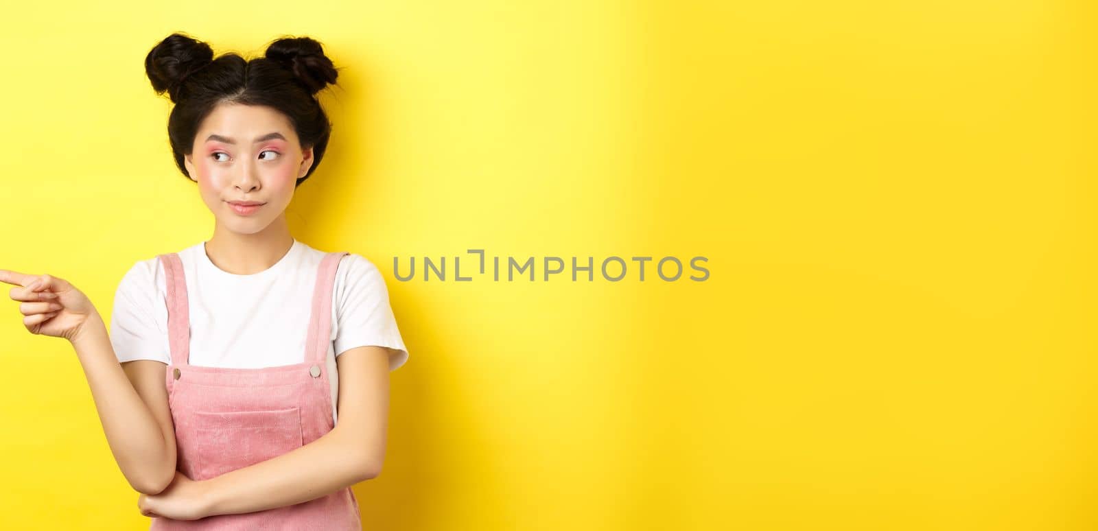 Cute beauty girl with makeup and summer clothes, pointing and looking left at promo banner, standing curious on yellow background.