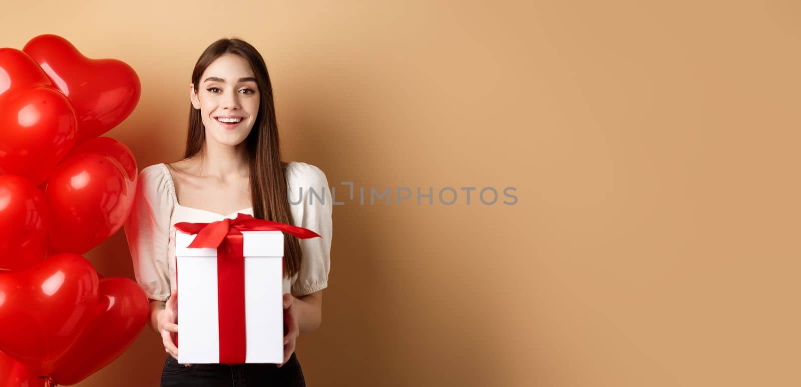 Romantic girl standing near heart balloons and holding surprise gift on Valentines day, smiling at camera happy, standing on beige background by Benzoix