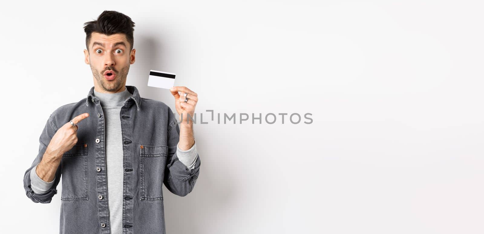 Wow look here. Excited guy pointing at plastic credit card and look amazed with awesome deal, standing against white background.