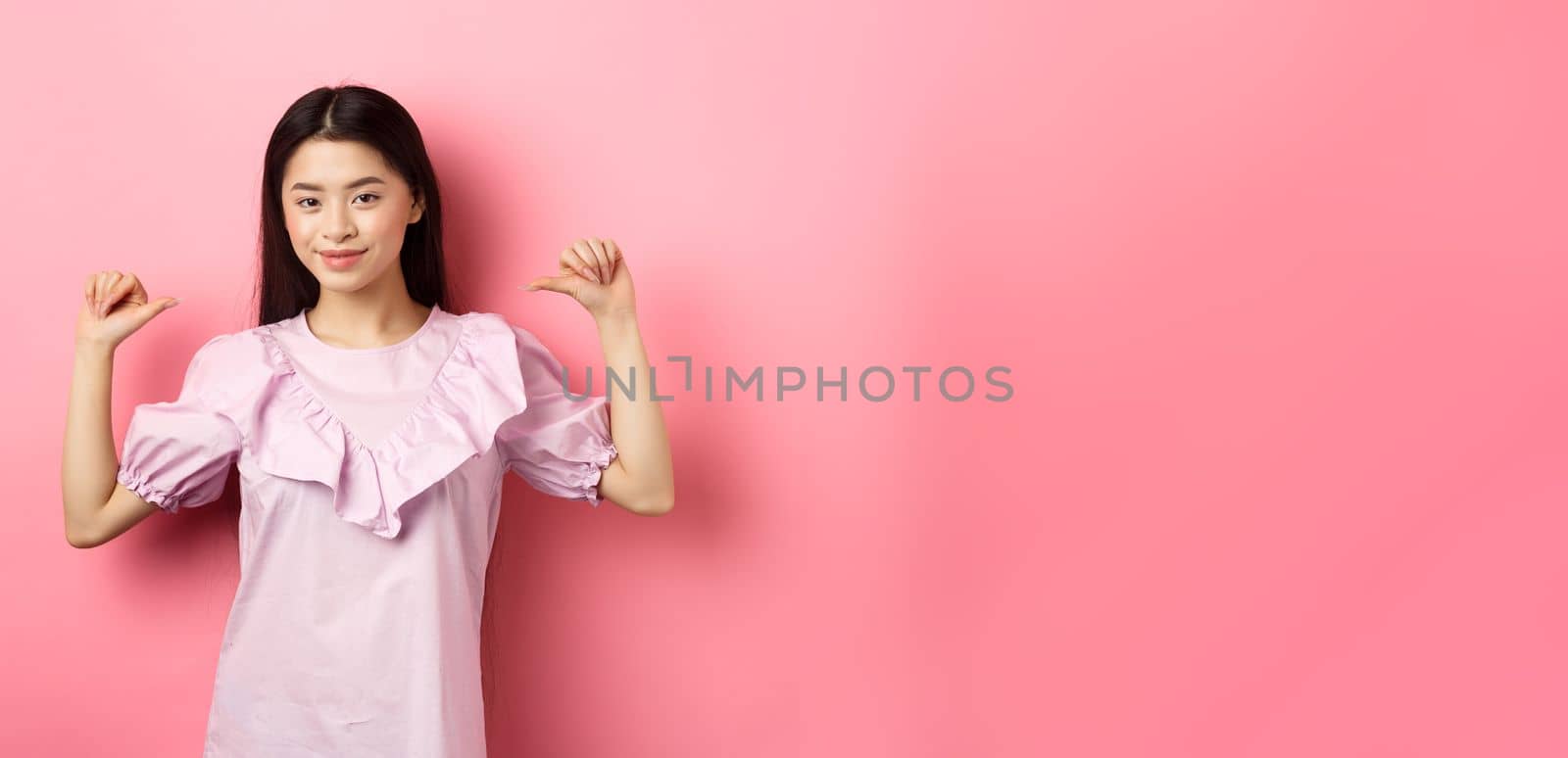 Confident asian girl smiling, pointing at herself, self-promoting personal achievements, standing in dress on pink background.