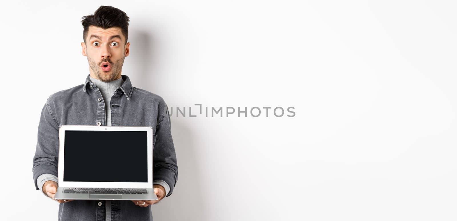Excited man demostrated empty laptop screen, say wow and look at camera amazed, checking out online offer, showing webpage, standing on white background.