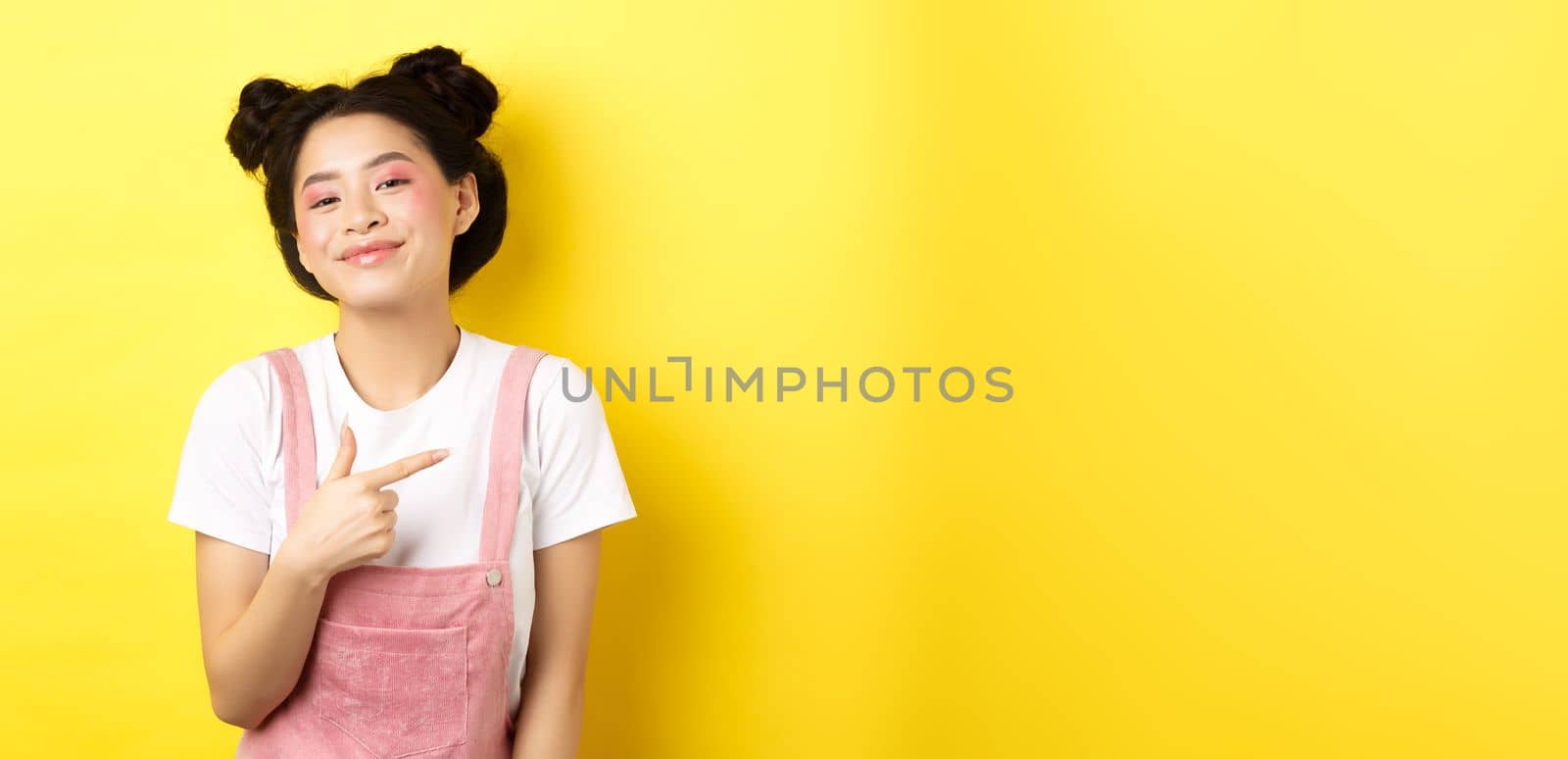 Stylish asian teen girl with romantic makeup, smiling cute and pointing finger right at logo, standing against yellow background.