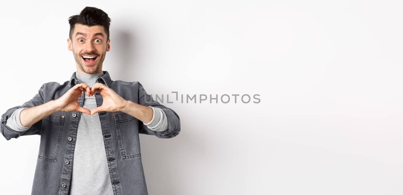 Romantic boyfriend showing heart gesture and looking with love at camera, celebrating White day after valentines, standing on white background. Romance concept.