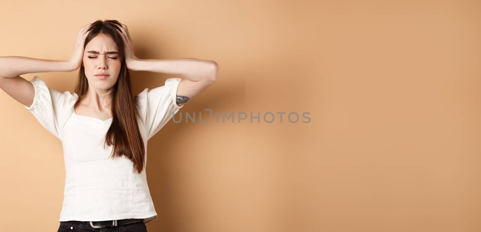 Frustrated young woman holding hands on head and feeling sad, standing distressed and troubled on beige background.