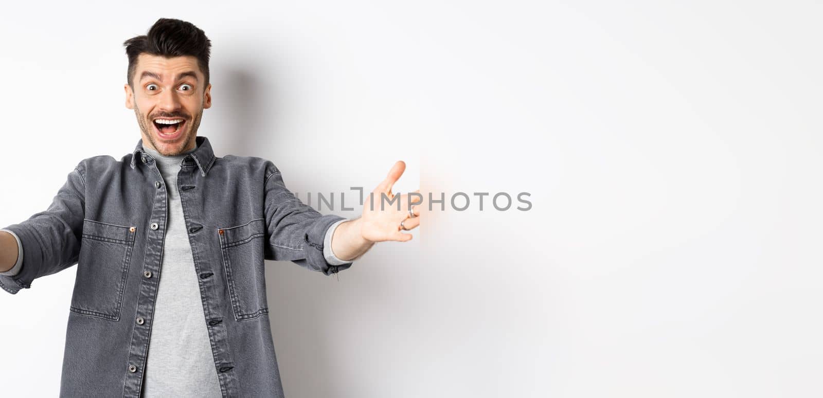 Excited young man stretch out hands for hug, cuddling you, welcoming guest and smiling, inviting people, standing on white background.