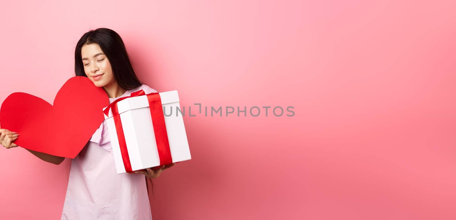 Valentines day concept. Romantic asian teen girl receive gifts on white day from lover, hugging big red heart card and present in box, smiling sensual, pink background.