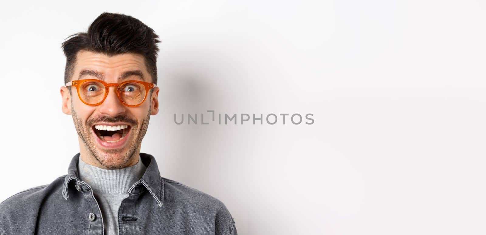 Excited funny man in glasses smiling, look with amazement and joy at awesome promo, standing happy against white background.