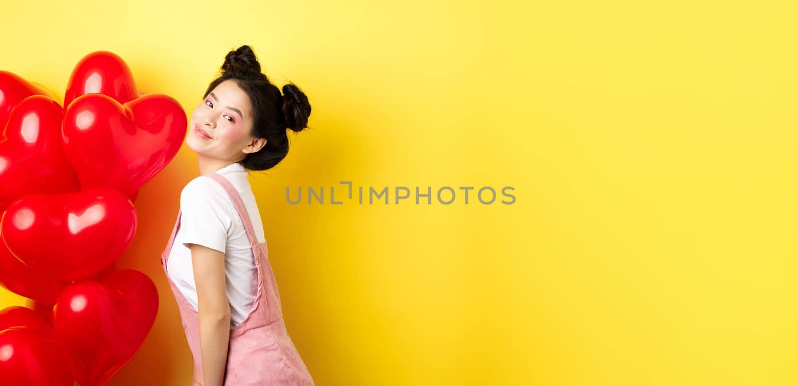 Valentines day and relationship concept. Fashionable asian woman posing near red hearts balloons, surprise for lover on special date, yellow background.