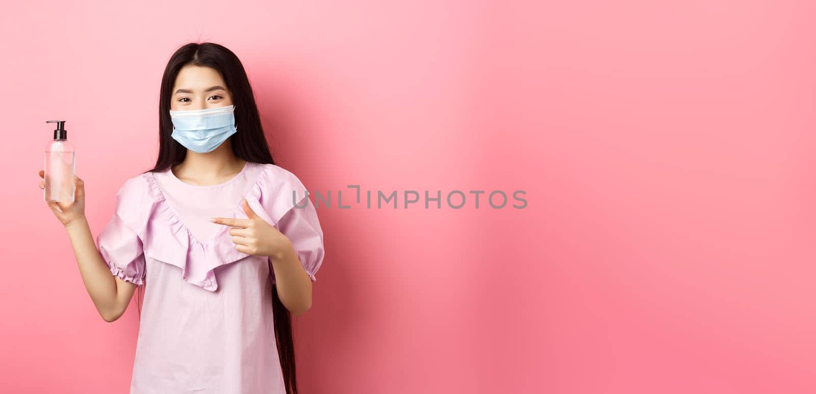 Healthy people and covid-19 pandemic concept. Cheerful asian woman in medical mask recommend hand sanitizer, pointing at antiseptic bottle, white background.