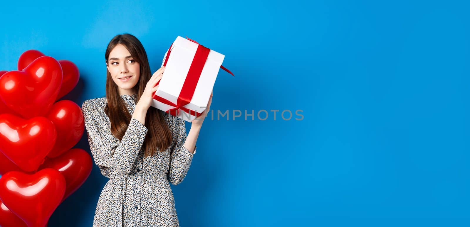 Valentines day. Beautiful woman shaking gift box to guess what inside, celebrating lovers holiday, standing near red hearts, blue background.