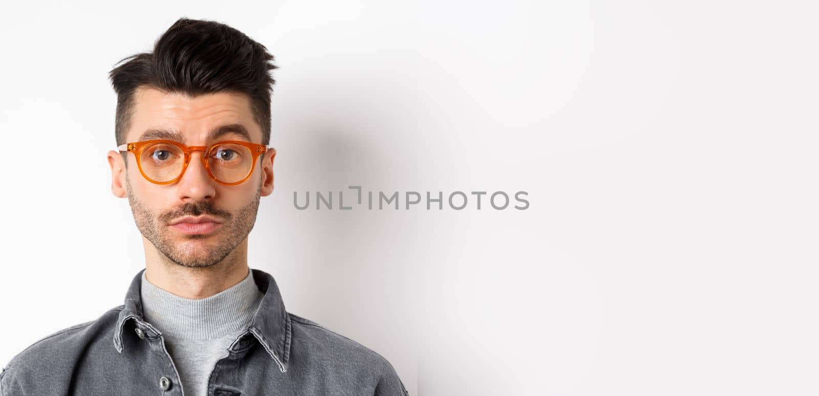 Attractive brunette man with moustache, wearing stylish glasses and look serious at camera, no emotion pokerface, standing against white background.