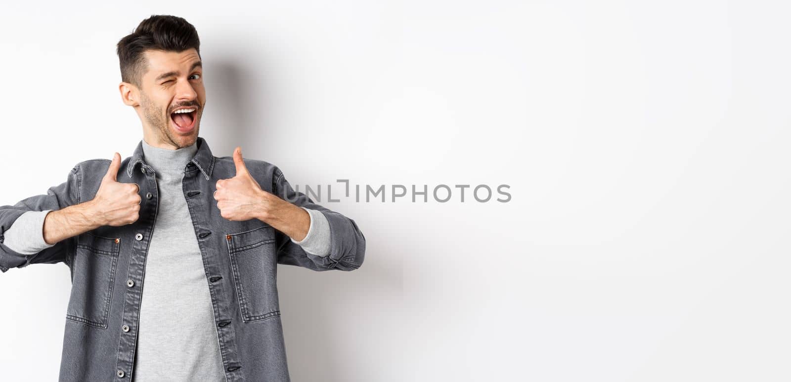 Happy young man with moustache winking and showing thumbs-up, recommending something, praise good thing, making compliment, standing on white background.