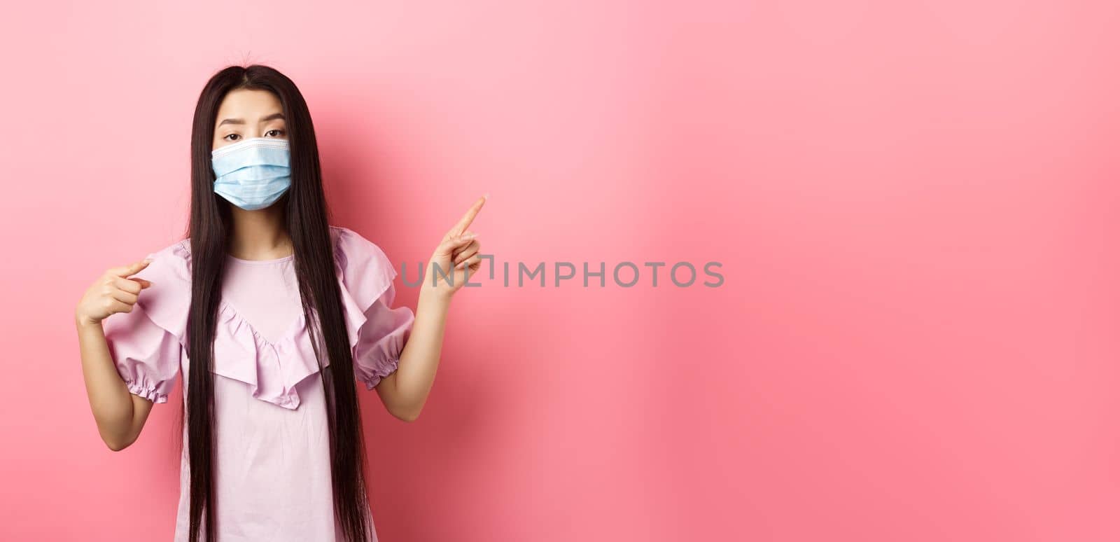 Healthy people and covid-19 pandemic concept. Bored asian woman in medical mask pointing right, showing logo, standing unamused on pink background by Benzoix