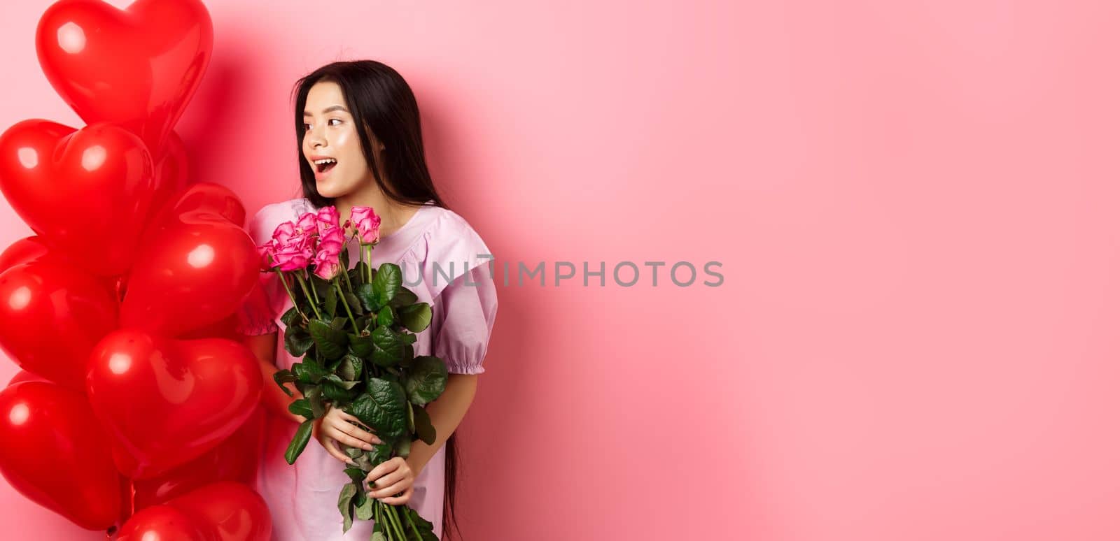 Portrait of asian teenage girl in love, holding flowers and looking at valentines day heart balloons, being on romantic date, pink background.