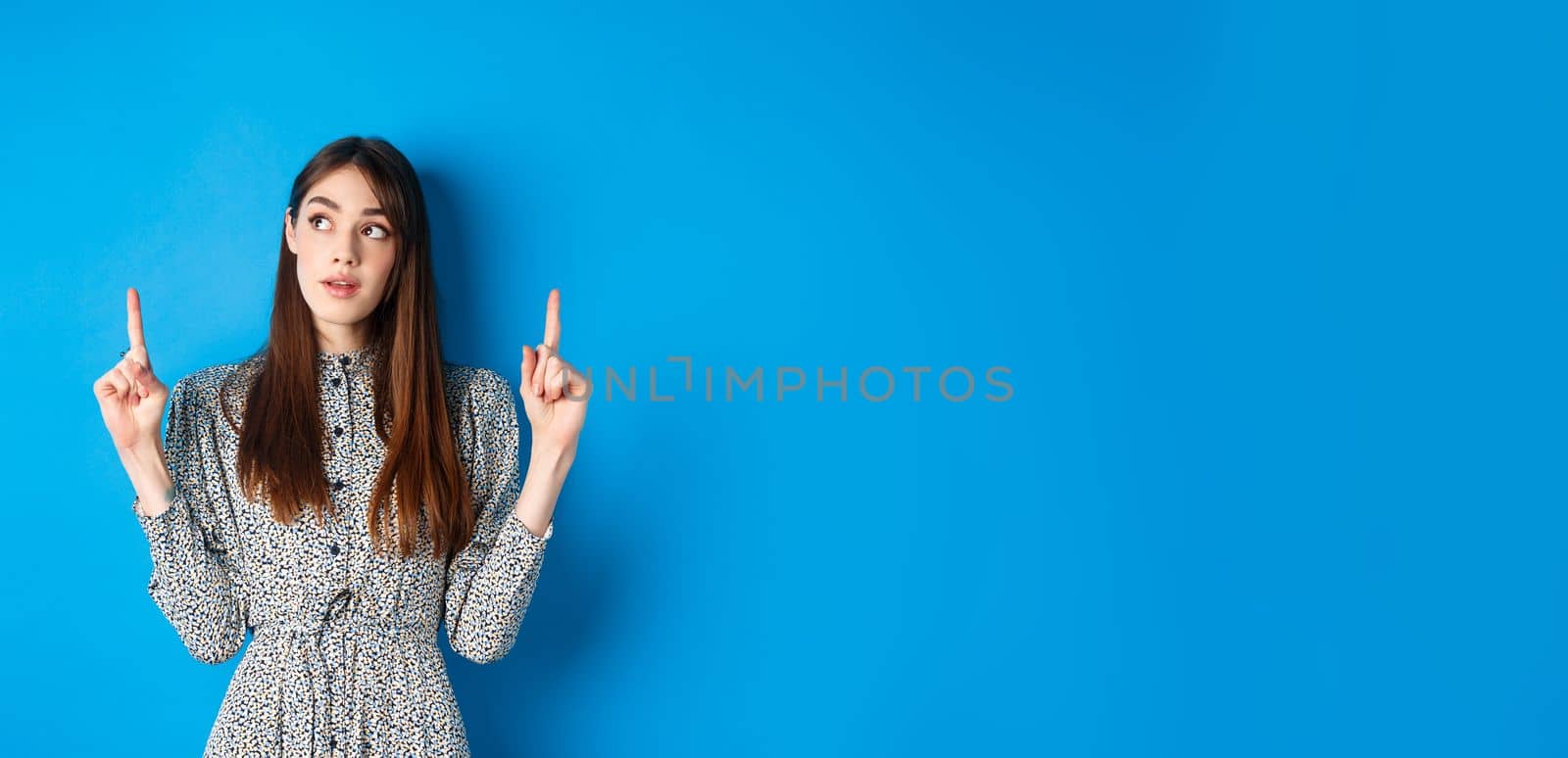 Pretty thoughtful woman pointing fingers up and thinking, standing pensive in dress on blue background.