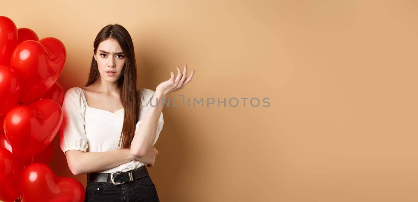 Valentines day and love concept. Confused girlfriend having argument with lover, raising hand and looking annoyed at camera, cant understand something, standing near hearts balloons.