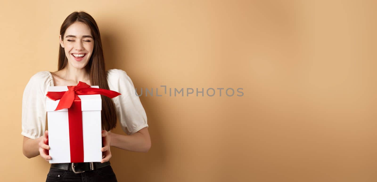 Happy smiling woman laughing, holding gift box from lover, enjoying romantic date on Valentines day, standing on beige background.