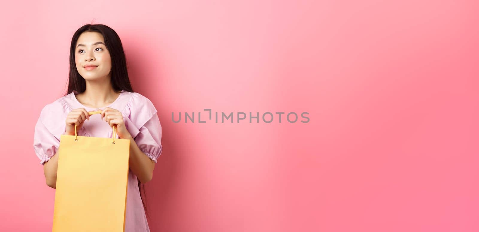 Beautiful asian girl dreaming and holding shopping bag, standing on pink background by Benzoix