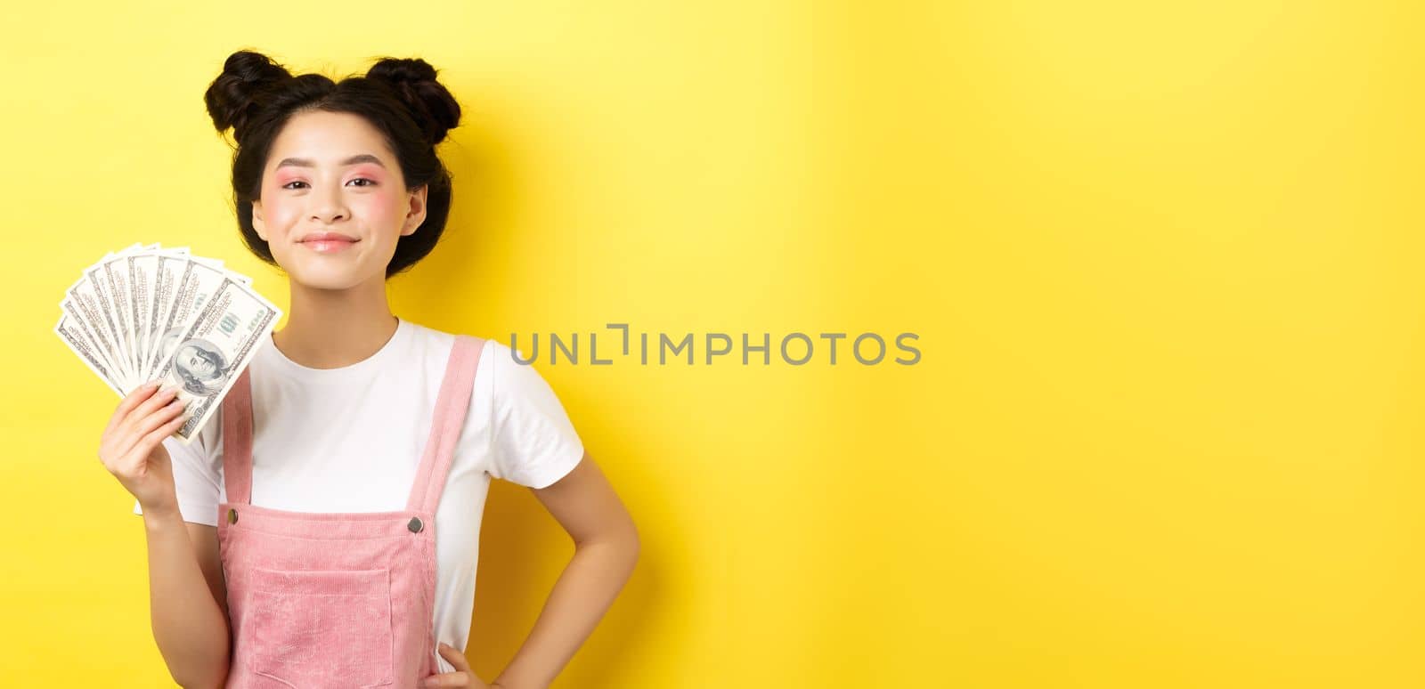 Shopping. Smiling asian girl with stylish makeup, showing dollar bills with pleased face, making money, standing on yellow background by Benzoix