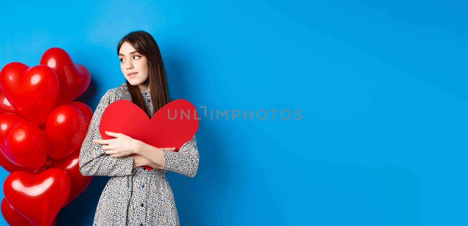 Valentines day. Beautiful and romantic woman looking pensive at balloons, hugging big red heart and smiling, waiting for love, blue background.