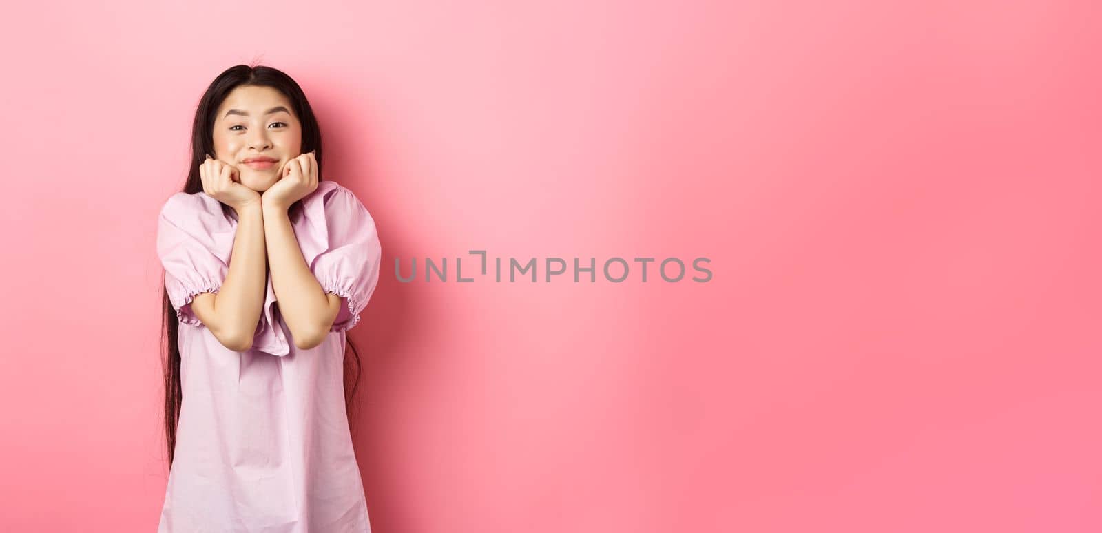 Cute and happy asian woman waiting for something with excitement, lean face on hands and watching lovely scene with admiration, standing against pink background.