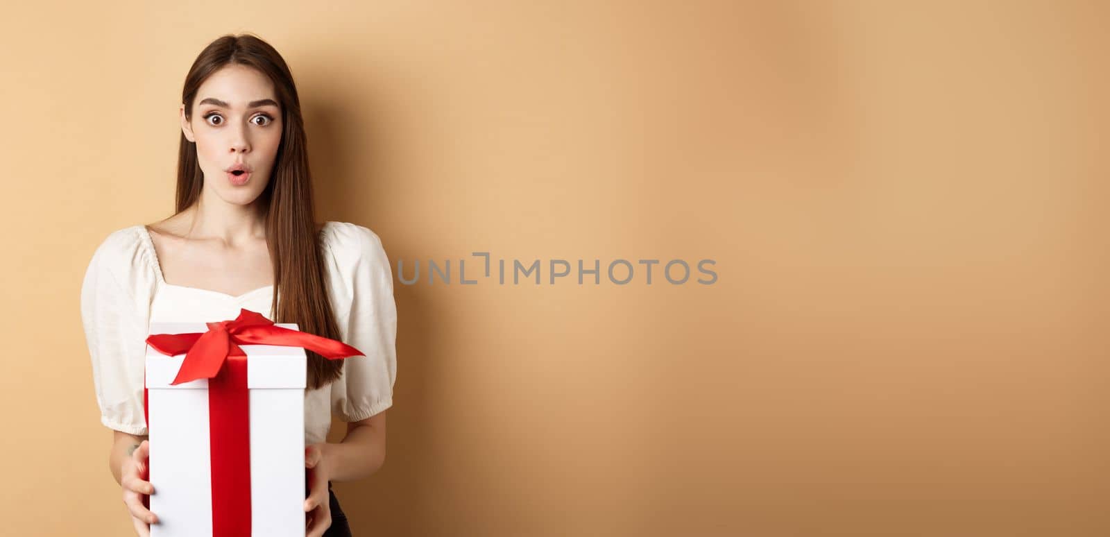 Image of surprised girl receive gift on Valentines day, looking with disbelief and holding present, standing on beige background.