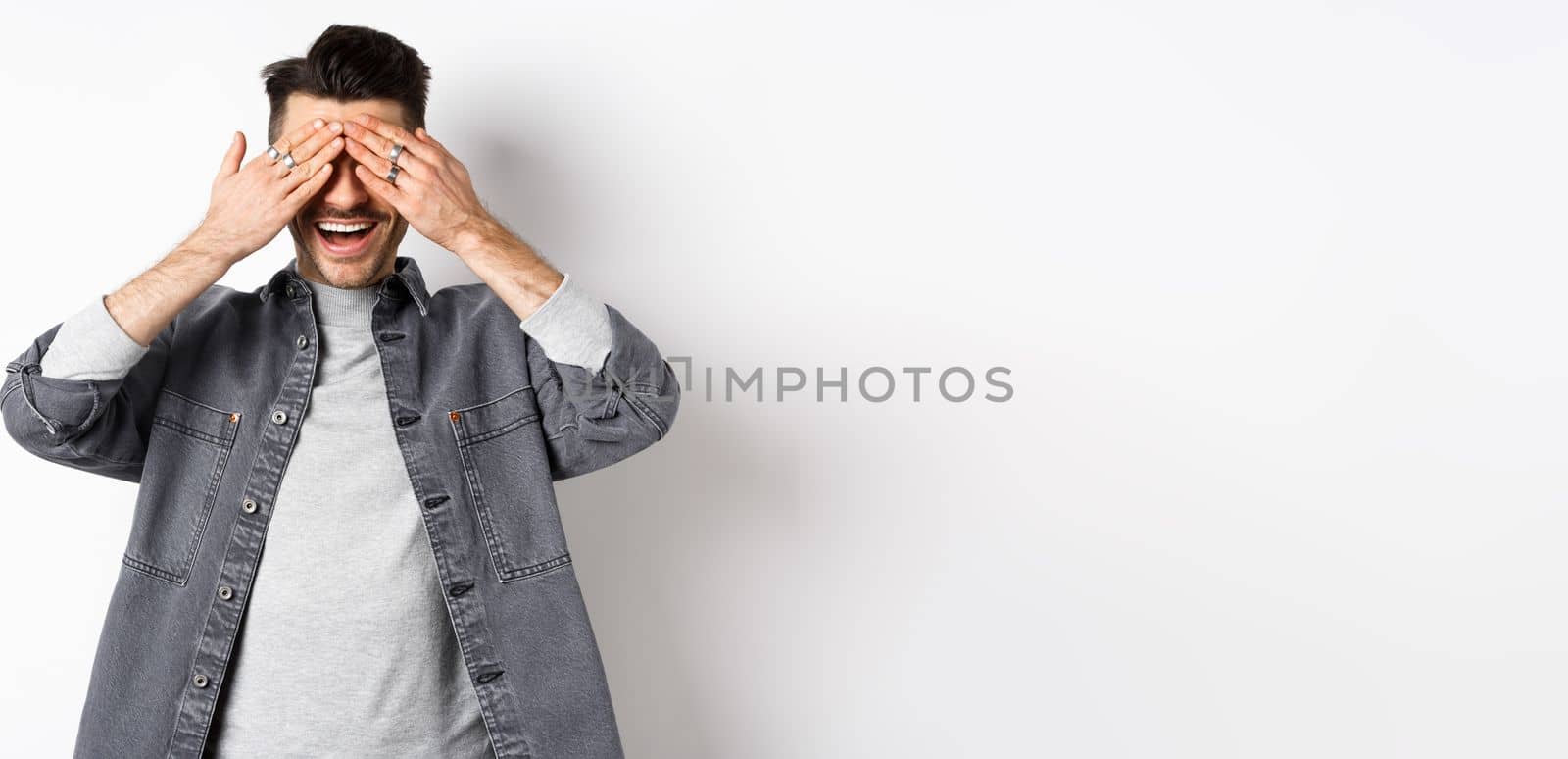 Handsome happy man cover his eyes and waiting for surprise, smiling excited, standing in casual clothes on white background.