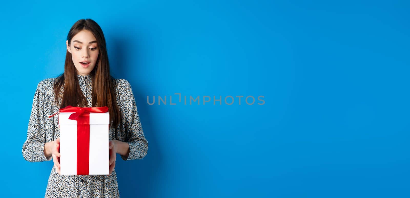 Holidays. Surprised young woman looking at gift with amazed face, receive Valentines day present, standing in dress on blue background by Benzoix