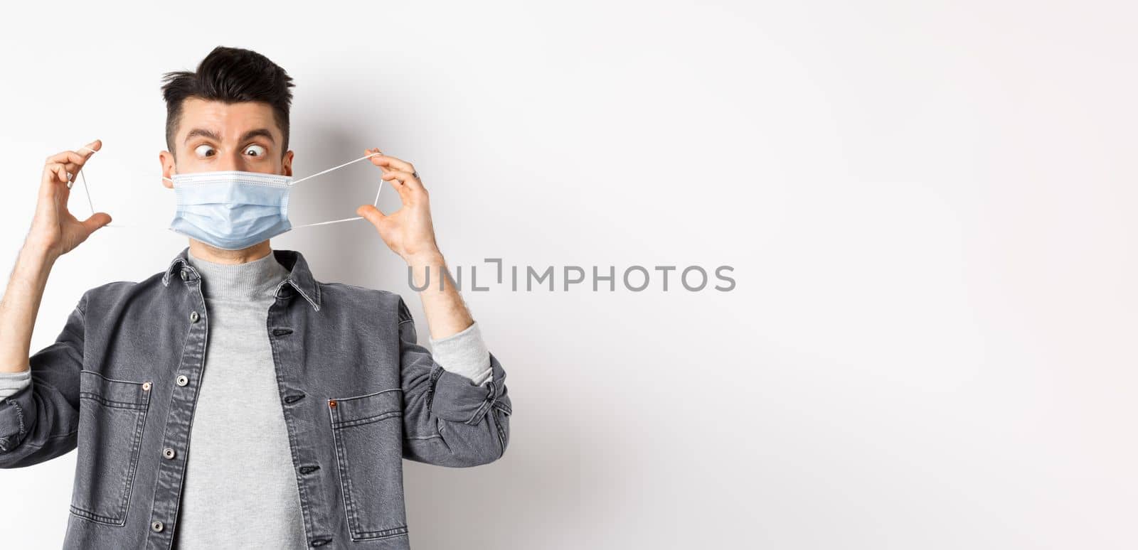 Health, covid and pandemic concept. Funny man squinting eyes at medical mask, playing with it, standing on white background.