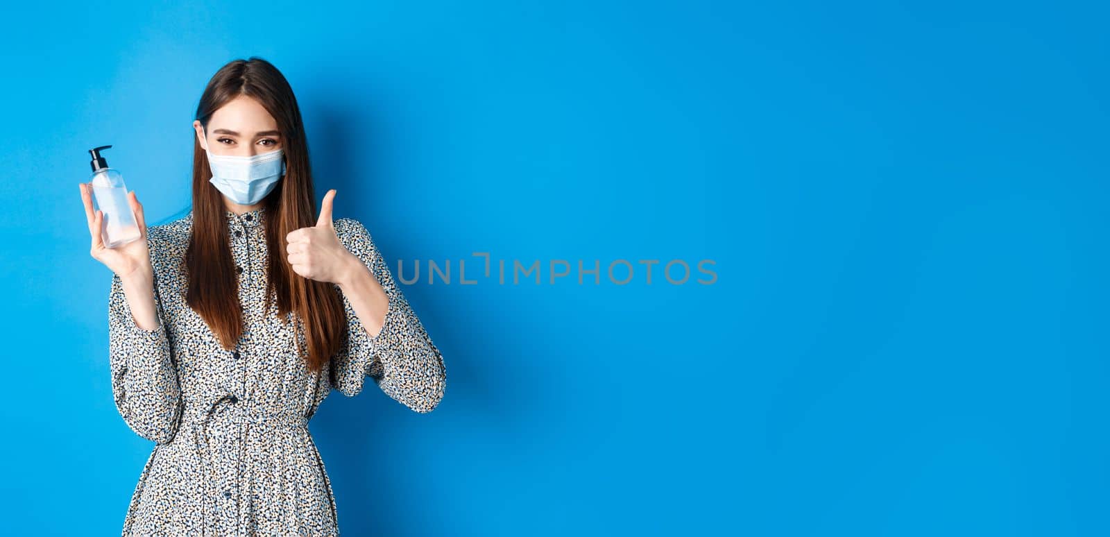 Covid-19, social distancing and healthcare concept. Beautiful girl in medical mask using hand saniziter, holding antiseptic bottle and showing thumbs up, standing on blue background by Benzoix