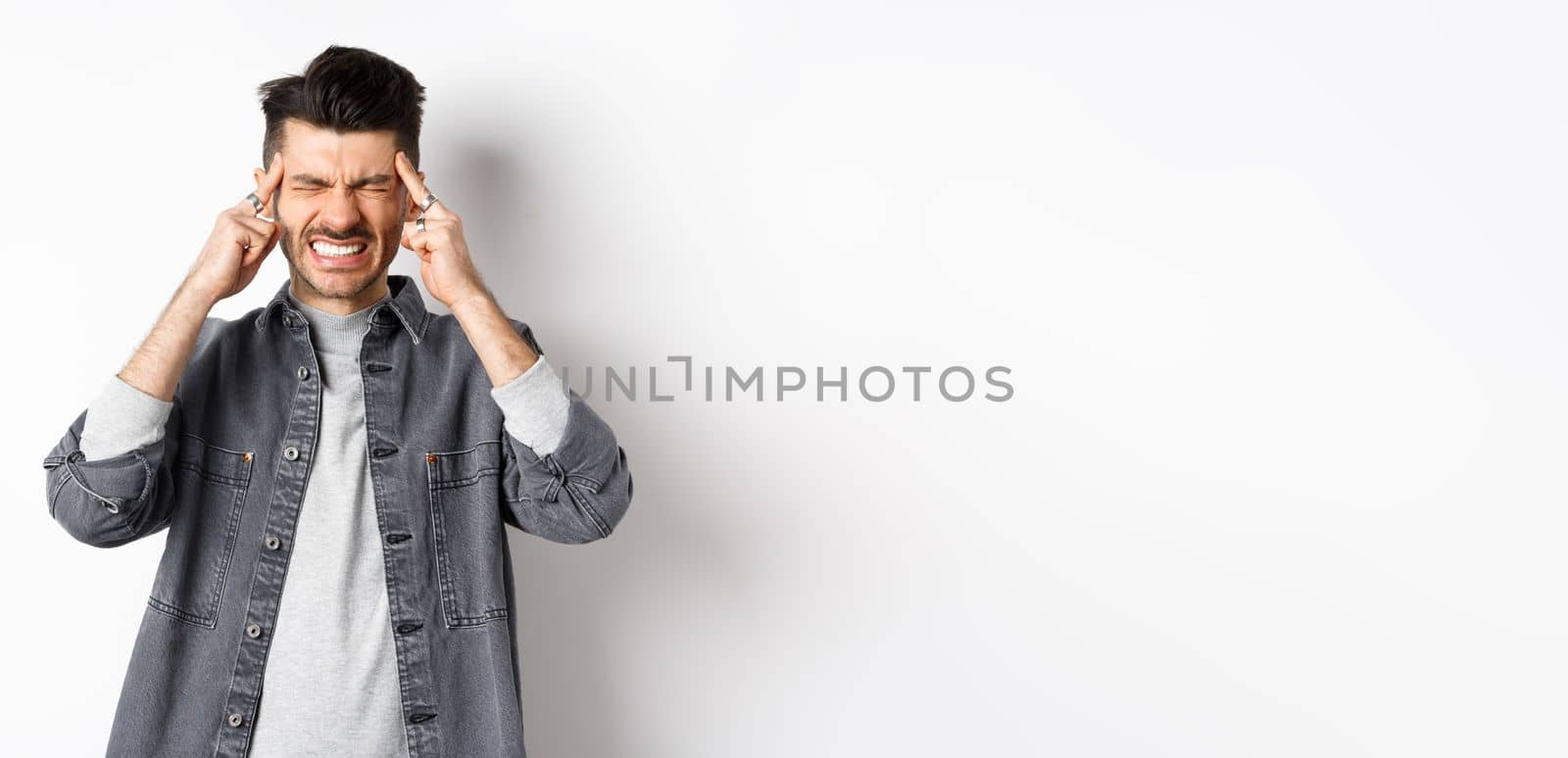 Man with headache grimacing and clenching teeth from painful migraine, touching head temples, suffering migraine, need painkillers, standing on white background.