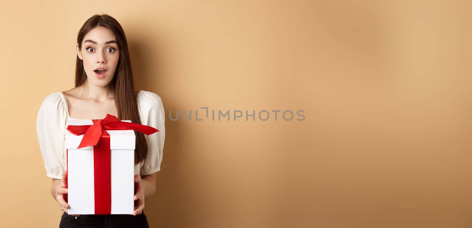 Amazed beautiful girl receiving gift from secret admirer on Valentines day, looking surprised at camera, holding box and standing on beige background.