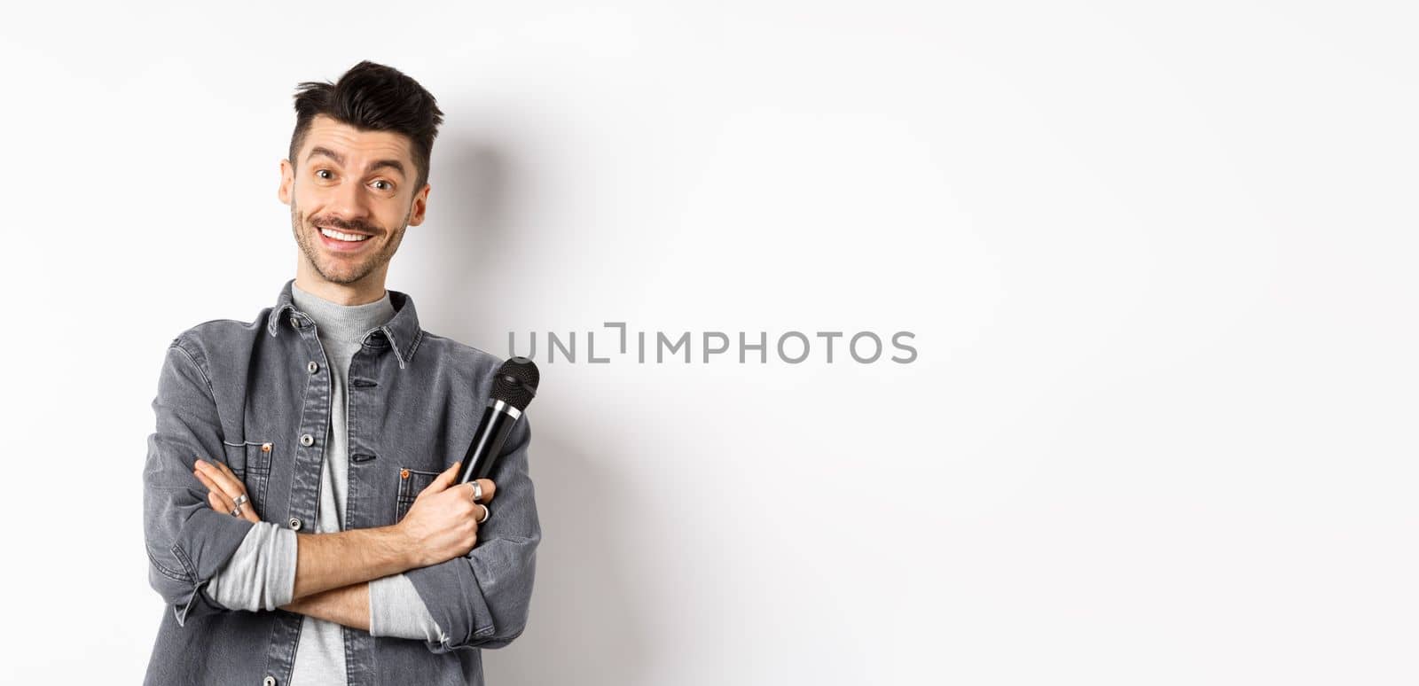 Handsome caucasian guy with moustache cross arms on chest, holding mic and smiling at camera, perform on stage with microphone, standing against white background.