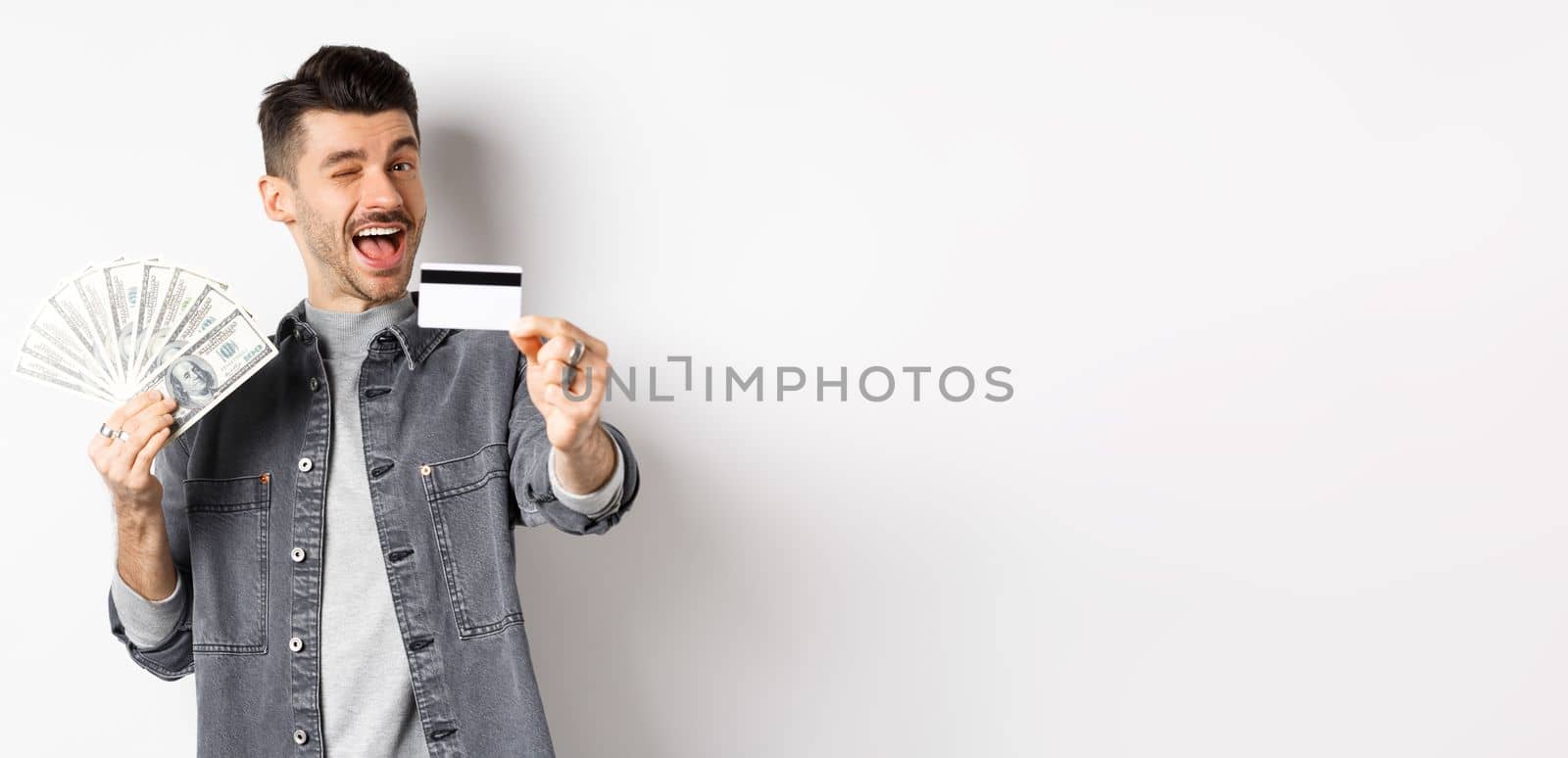 Image of handsome guy holding dollar bills but suggesting use plastic credit card, smiling friendly at camera, standing on white background.