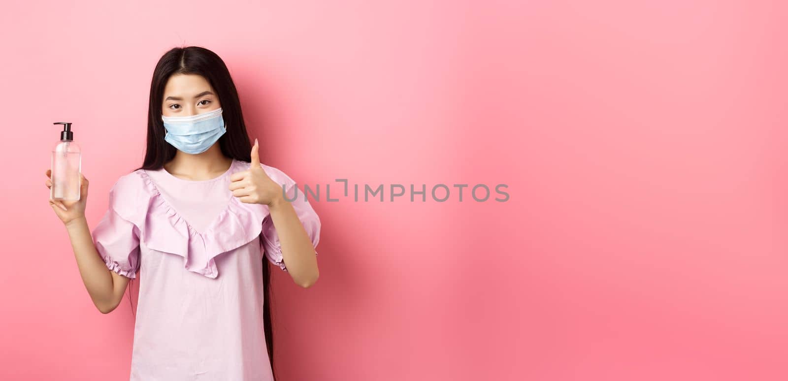 Healthy people and covid-19 pandemic concept. Cheerful asian woman in medical mask recommend hand sanitizer, showing thumb-up, praise antiseptic bottle, white background.