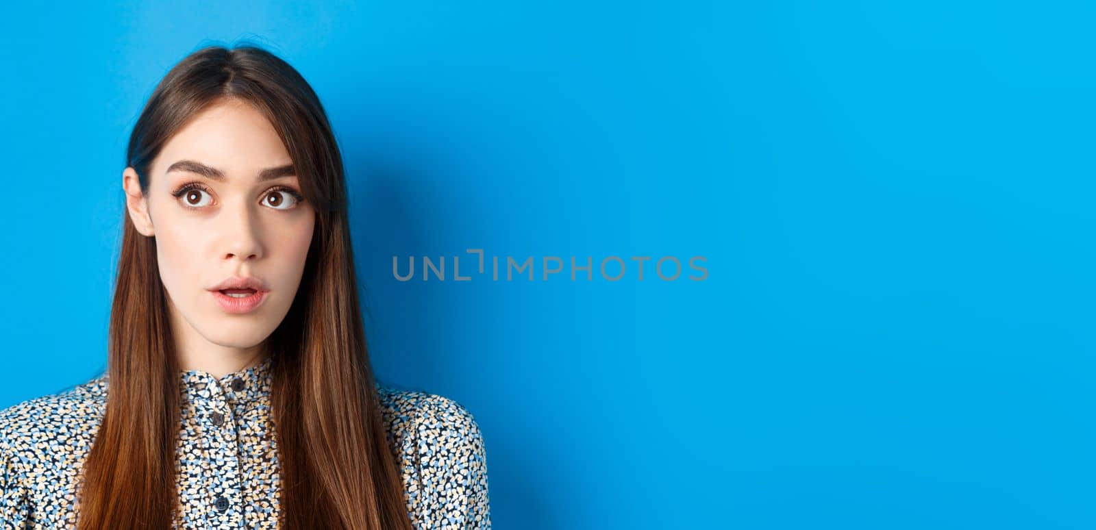 Close-up portrait of surprised young woman with natural beauty, looking at upper left corner and gasping, standing on blue background.