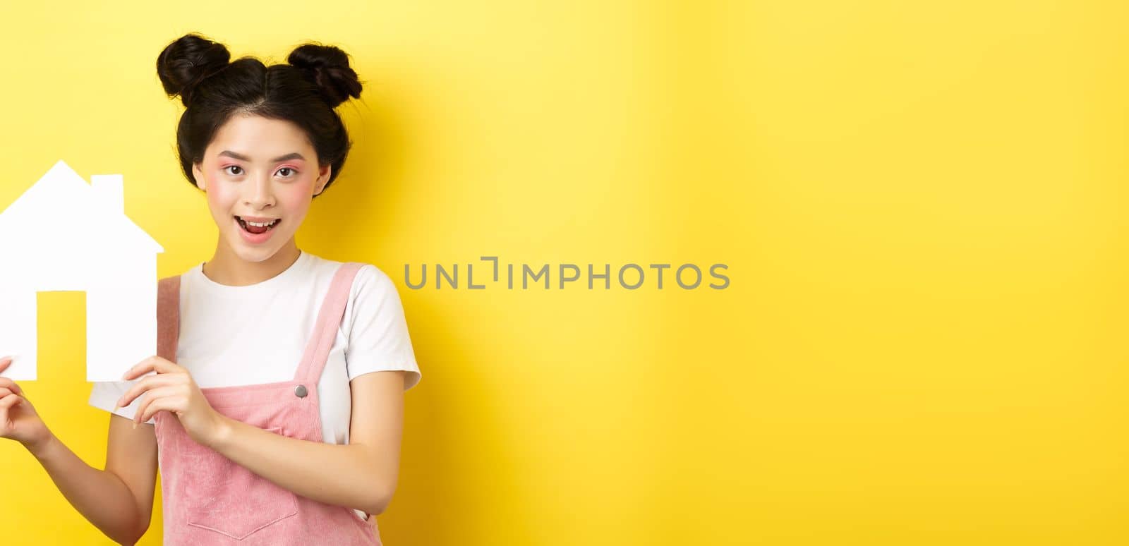 Real estate and family concept. Cute asian woman with bright makeup and stylish hairbuns, showing paper house cutout, smiling determined, yellow background by Benzoix