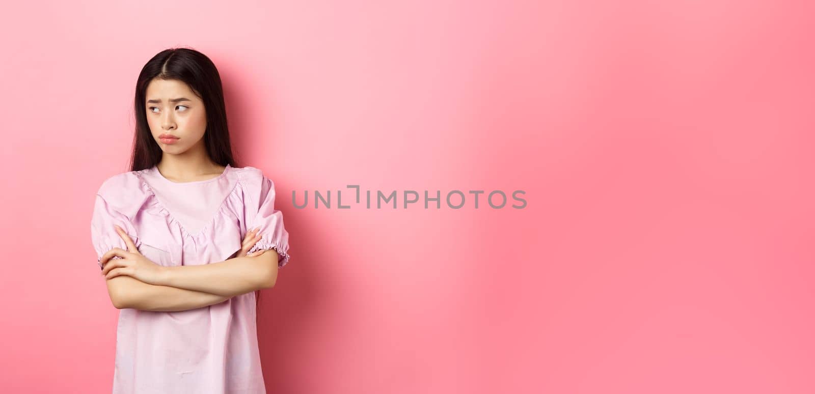 Sad and gloomy asian woman look away at logo, sulking from unfair situation, cross arms on chest disappointed, standing against pink background.