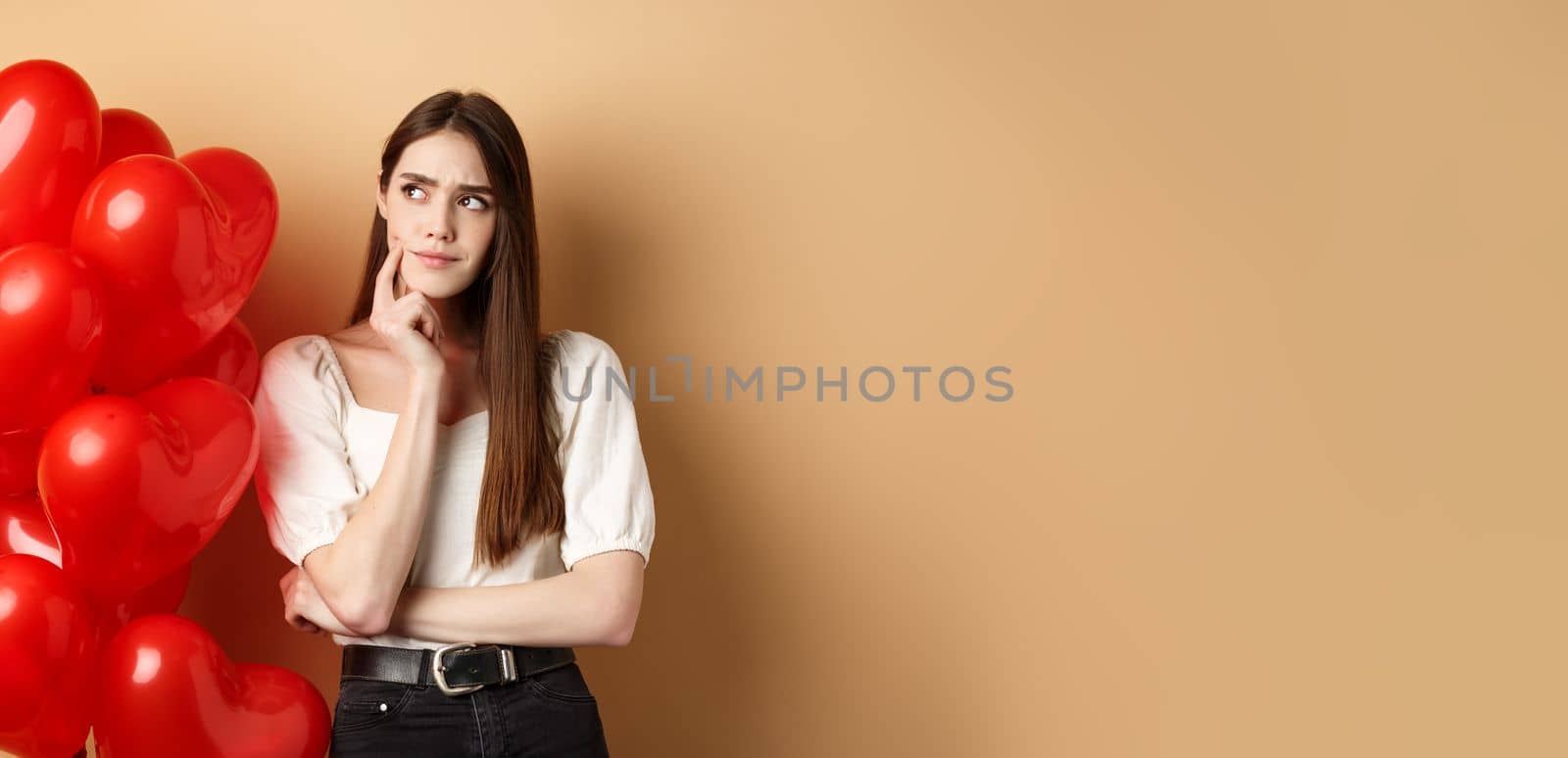 Valentines day and love concept. Pensive woman in trendy clothes and makeup, thinking and frowning, looking aside doubtful, standing near heart balloons, beige background by Benzoix