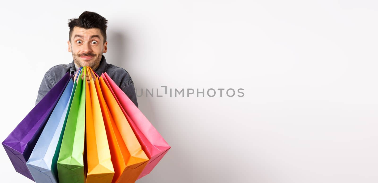 Excited young man carry colorful shopping bags and smiling happy, shopper buying on sale, standing on white background.