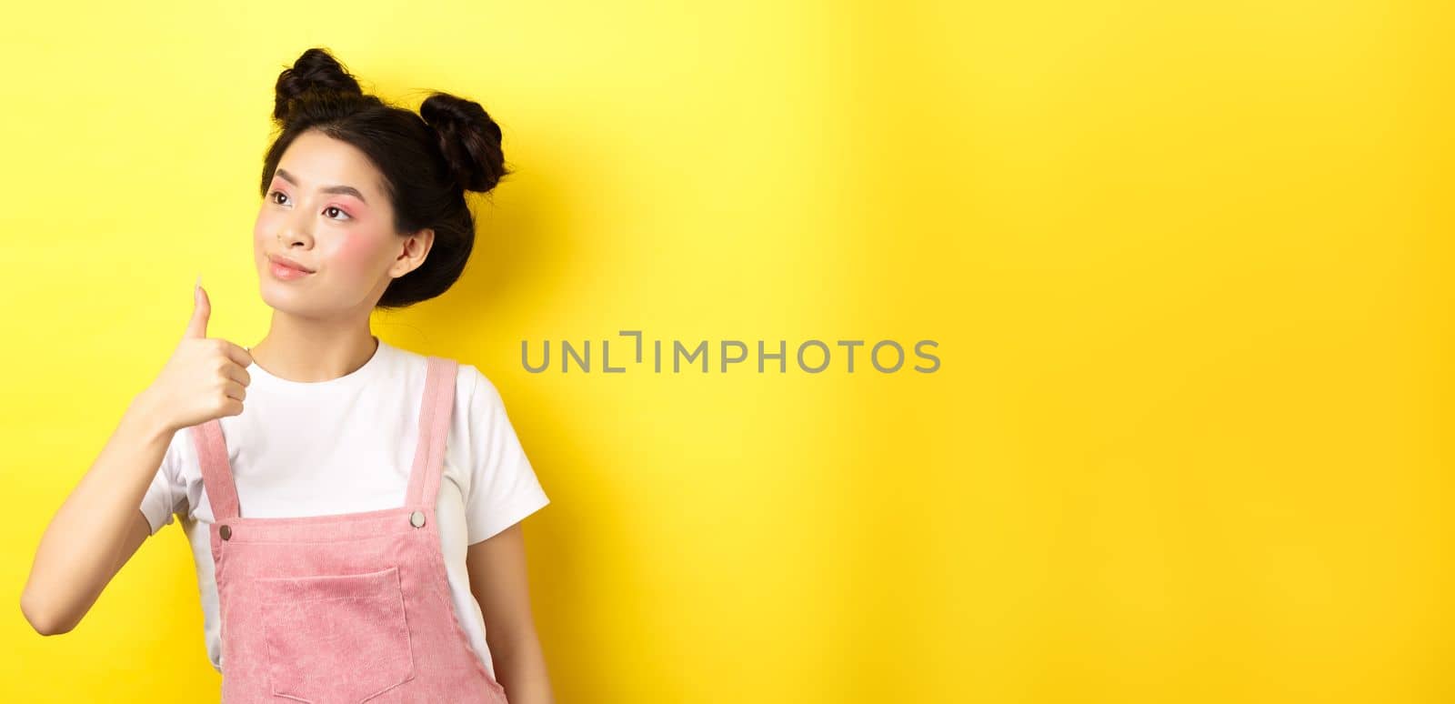 Glamour asian girl with pink makeup and summer clothes, looking left at logo with thumb up, approve and praise, standing on yellow background by Benzoix